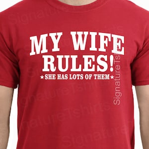 May include: Red t-shirt with white text that reads "MY WIFE RULES!" and "SHE HAS LOTS OF THEM" with a star on either side of the text.