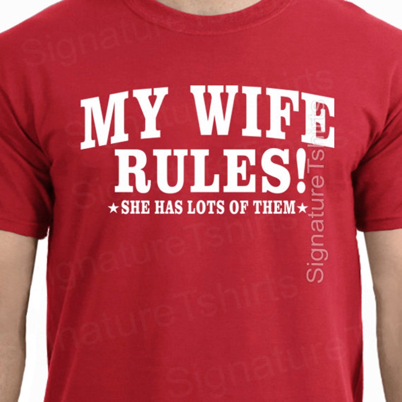 May include: Red t-shirt with white text that reads "MY WIFE RULES!" and "SHE HAS LOTS OF THEM" with a star on either side of the text.