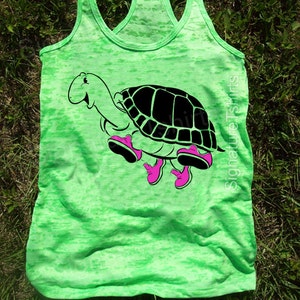 May include: A green tank top with a black and white cartoon turtle wearing pink shoes. The text "Signature Tshirts" is printed on the right side of the tank top.