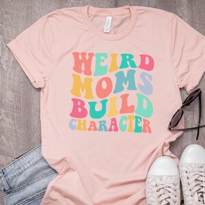 Pode incluir: Camiseta rosa com o texto "Weird Moms Build Character" em fonte retr&ocirc; colorida.