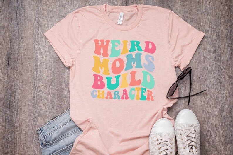 Pode incluir: Camiseta rosa com o texto "Weird Moms Build Character" em fonte retr&ocirc; colorida.