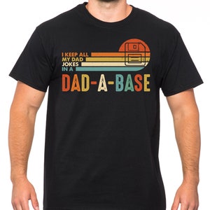 Op de afbeelding: Zwart T-shirt met een retro ontwerp met een floppy disk afbeelding en de tekst "I KEEP ALL MY DAD JOKES IN A DAD-A-BASE" in oranje, geel en blauw. Het shirt heeft een ronde hals en korte mouwen.