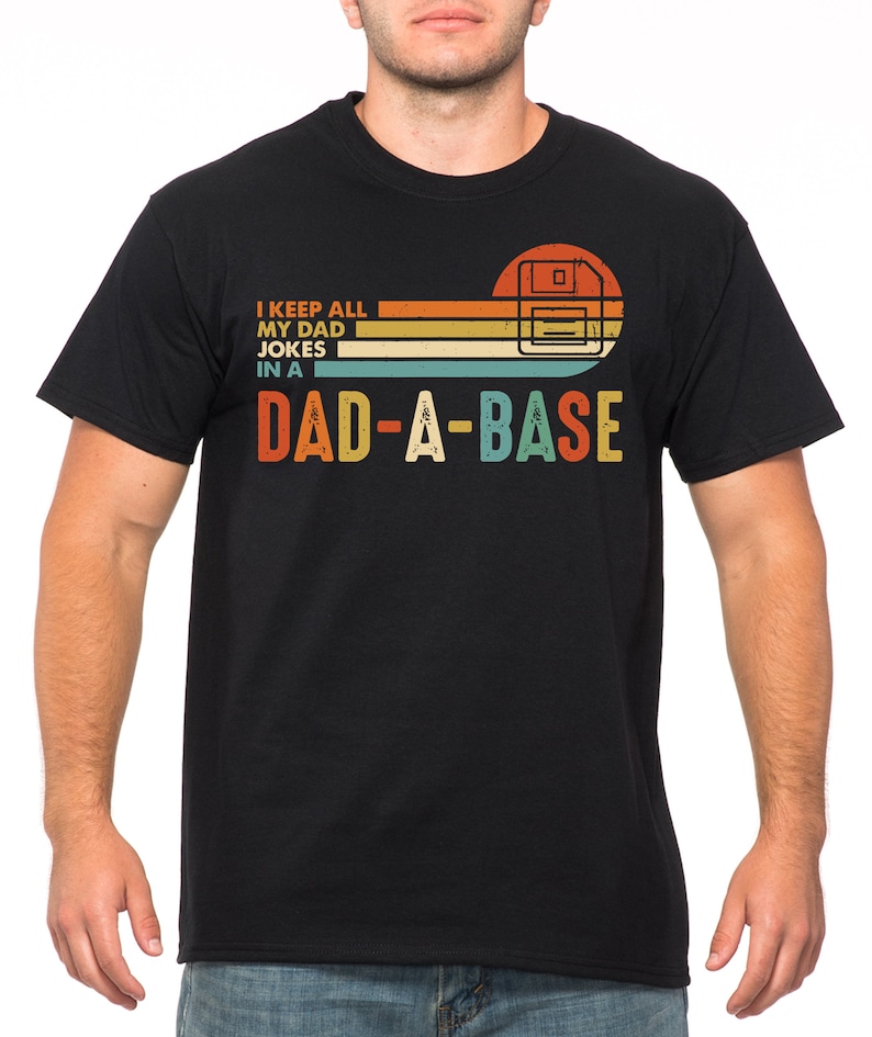 Op de afbeelding: Zwart T-shirt met een retro ontwerp met een floppy disk afbeelding en de tekst "I KEEP ALL MY DAD JOKES IN A DAD-A-BASE" in oranje, geel en blauw. Het shirt heeft een ronde hals en korte mouwen.
