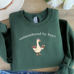 May include: A dark green sweatshirt with the text "outnumbered by boys" in white. The sweatshirt features a cartoon goose wearing a hat and holding a football. The goose is also holding a tray of food.