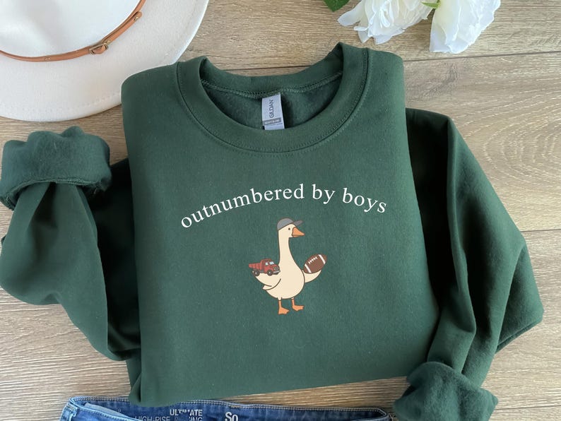 May include: A dark green sweatshirt with the text "outnumbered by boys" in white. The sweatshirt features a cartoon goose wearing a hat and holding a football. The goose is also holding a tray of food.