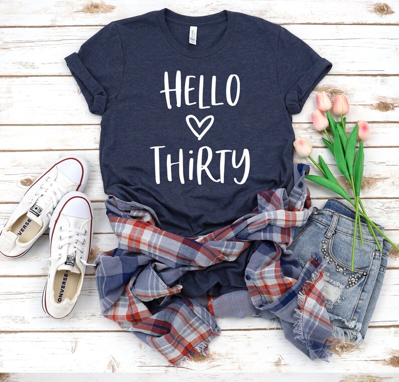May include: A navy blue t-shirt with the text "Hello Thirty" in white with a heart symbol between the words. The shirt is laid out on a white wooden surface with a plaid shirt, denim shorts, and pink flowers.