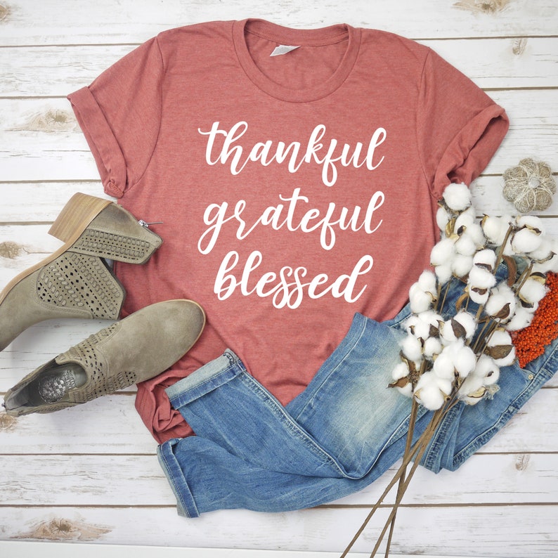 May include: A rust-colored t-shirt with the words "thankful grateful blessed" printed in white on the front. The shirt is laid flat on a white wooden surface with a pair of brown ankle boots and a bunch of white cotton stems.