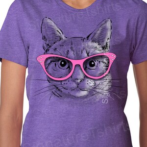 May include: A purple t-shirt with a graphic of a cat wearing pink glasses. The cat's eyes are looking directly at the viewer.