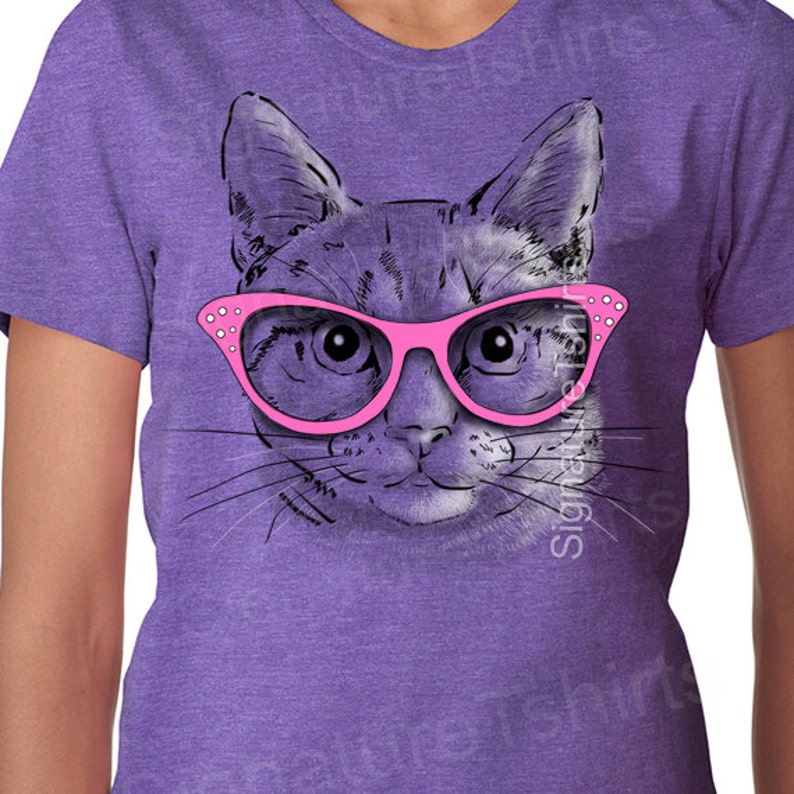 May include: A purple t-shirt with a graphic of a cat wearing pink glasses. The cat's eyes are looking directly at the viewer.