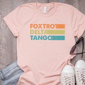 以下が含まれることがあります： レトロなスタイルでカラフルなストライプが入った「FOXTROT DELTA TANGO」の文字がプリントされたピンクのTシャツです。