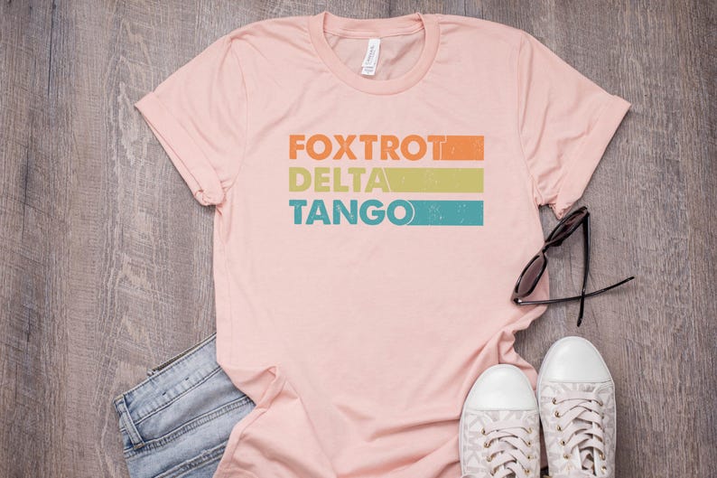 以下が含まれることがあります： レトロなスタイルでカラフルなストライプが入った「FOXTROT DELTA TANGO」の文字がプリントされたピンクのTシャツです。