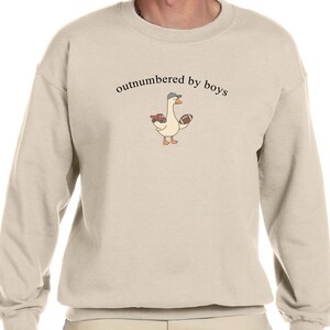 May include: A beige sweatshirt with the phrase "outnumbered by boys" printed in black. The sweatshirt features a cartoon goose wearing a hat and holding a football and a red truck.