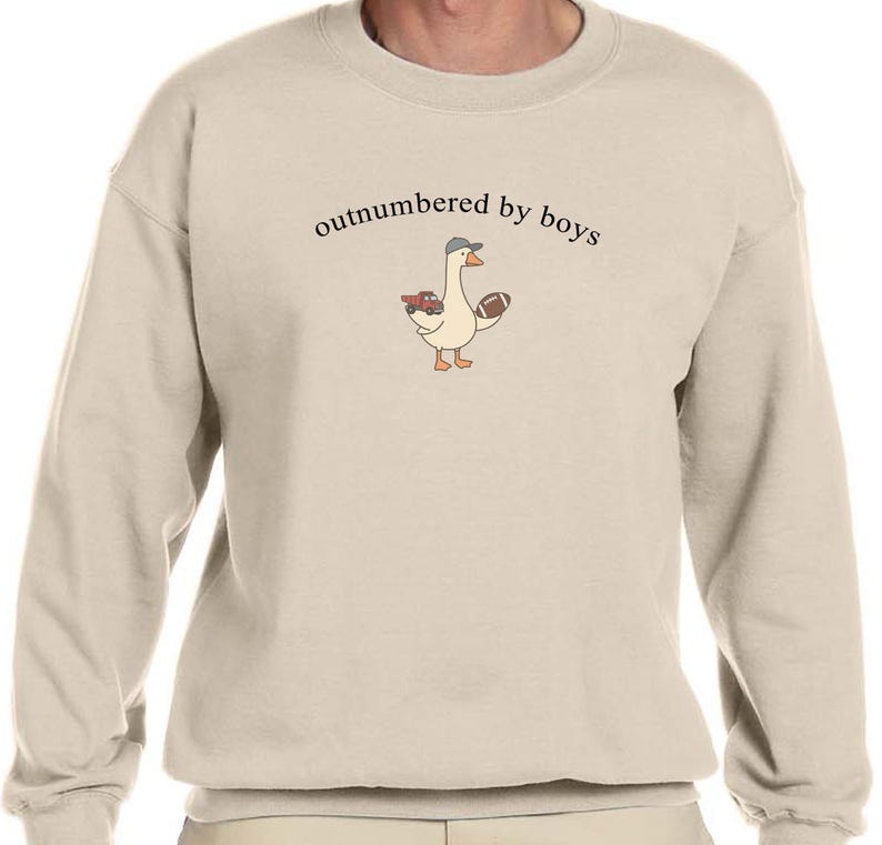 May include: A beige sweatshirt with the phrase "outnumbered by boys" printed in black. The sweatshirt features a cartoon goose wearing a hat and holding a football and a red truck.