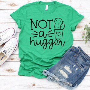 May include: Green t-shirt with the text "NOT a hugger" and a black and white illustration of a cactus in a pot with a heart.