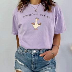 May include: A lilac t-shirt featuring the phrase "outnumbered by boys" and a cartoon goose wearing a cap, holding a football and a toy truck. The shirt has a relaxed fit and short sleeves.