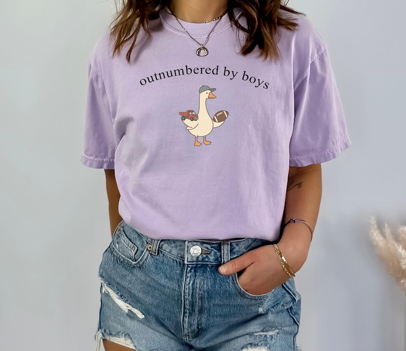 May include: A lilac t-shirt featuring the phrase "outnumbered by boys" and a cartoon goose wearing a cap, holding a football and a toy truck. The shirt has a relaxed fit and short sleeves.