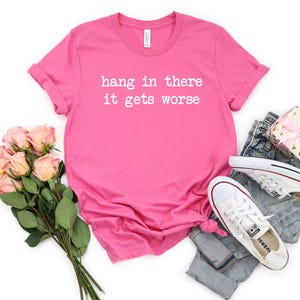 May include: A bright pink t-shirt with the phrase "hang in there it gets worse" printed in white. The shirt is styled with a pair of blue jeans, white sneakers, a bouquet of pink roses, and a small gift box.
