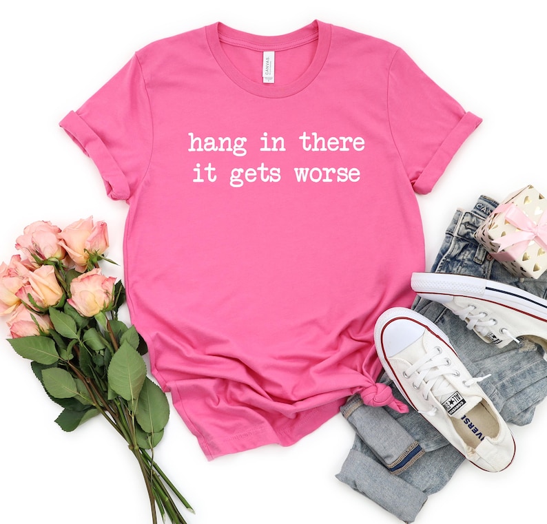 May include: A bright pink t-shirt with the phrase "hang in there it gets worse" printed in white. The shirt is styled with a pair of blue jeans, white sneakers, a bouquet of pink roses, and a small gift box.