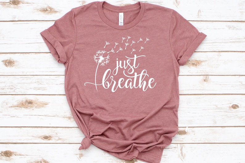 Op de afbeelding: Een mauve t-shirt met een witte paardenbloem afbeelding en de tekst "just breathe" in een cursief schrift.