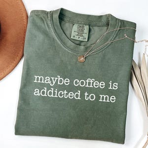 May include: An olive green t-shirt with the phrase "maybe coffee is addicted to me" printed in white. A gold necklace with a pendant and a brown hat are also in the image. The shirt is folded neatly.