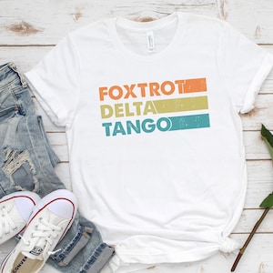 以下が含まれることがあります： オレンジ、イエロー、ブルーのストライプでレトロなスタイルで「FOXTROT DELTA TANGO」とプリントされた白いTシャツです。