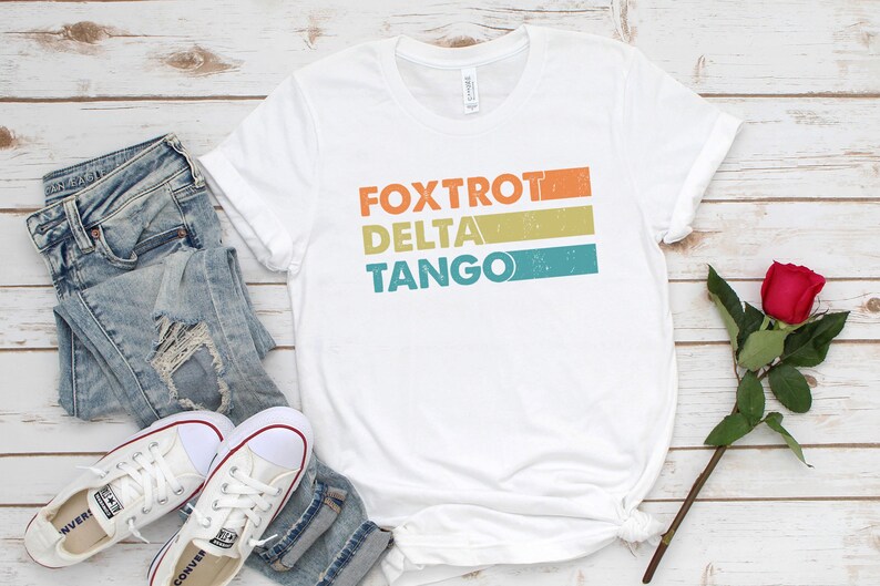以下が含まれることがあります： オレンジ、イエロー、ブルーのストライプでレトロなスタイルで「FOXTROT DELTA TANGO」とプリントされた白いTシャツです。