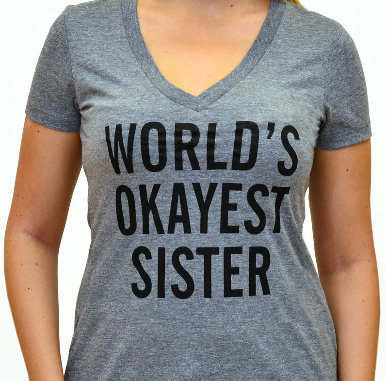 May include: A heather gray v-neck t-shirt with the text "WORLD'S OKAYEST SISTER" printed in black. The shirt is made of a soft material and has short sleeves. The text is centered on the front of the shirt.