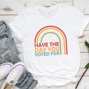 May include: White t-shirt with a rainbow graphic and the text "Have the day you voted for." The shirt is paired with light-wash denim jeans and white sneakers. A red rose is placed on the wooden surface.