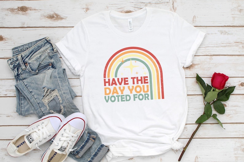 May include: White t-shirt with a rainbow graphic and the text "Have the day you voted for." The shirt is paired with light-wash denim jeans and white sneakers. A red rose is placed on the wooden surface.