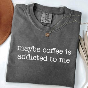 May include: A charcoal gray t-shirt with the phrase "maybe coffee is addicted to me" in white lettering. A gold-colored necklace with a pendant rests on the shirt. The shirt is folded, and a brown hat is partially visible.