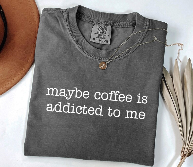 May include: A charcoal gray t-shirt with the phrase "maybe coffee is addicted to me" in white lettering. A gold-colored necklace with a pendant rests on the shirt. The shirt is folded, and a brown hat is partially visible.