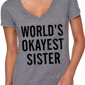 May include: A heather gray V-neck t-shirt with the text "WORLD'S OKAYEST SISTER" in bold black letters. The shirt is a casual style, suitable for everyday wear. The text is centered on the front of the shirt.