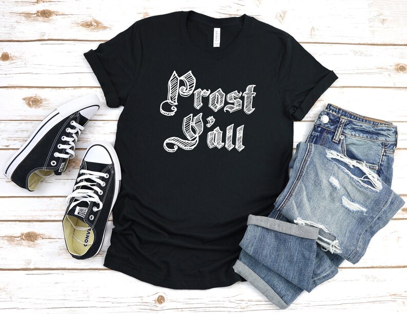 May include: Black t-shirt with the text "Prost Y'all" in white, stylized lettering. The shirt is paired with blue denim jeans and black canvas sneakers with white laces. A casual, everyday outfit.