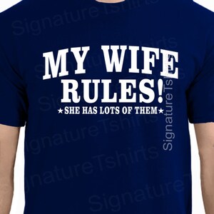 May include: A navy blue t-shirt with white text that reads "My Wife Rules!" and a smaller line of text that reads "She has lots of them."  The text is in a bold, blocky font.