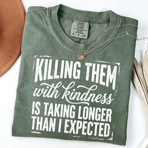 Puede incluir: Una camiseta de manga corta color verde oliva con cuello redondo. La prenda presenta un texto impreso en blanco que dice: "KILLING THEM with kindness IS TAKING LONGER THAN I EXPECTED," enmarcado por un borde rectangular desgastado.