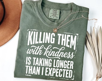 May include: An olive green short-sleeve t-shirt with a crew neckline. The shirt features white printed text that reads, "KILLING THEM with kindness IS TAKING LONGER THAN I EXPECTED," framed by a distressed rectangular border.