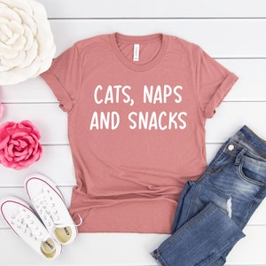 May include: A dusty rose colored t-shirt with the text "CATS, NAPS AND SNACKS" in white lettering. The t-shirt is laid flat on a white wooden surface with a pair of white sneakers, a pair of blue jeans, and three paper flowers.