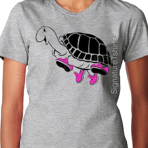 May include: A gray t-shirt with a black and white cartoon turtle wearing pink shoes. The turtle is walking and has a happy expression. The text "Signature Tshirts" is on the right side of the shirt.