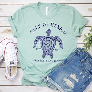 Puede incluir: Una camiseta verde claro con un dise&ntilde;o de tortuga marina azul marino y el texto "GULF OF MEXICO" y "STAY SALTY AND RESIST". La camiseta se combina con pantalones cortos de mezclilla y zapatillas blancas.