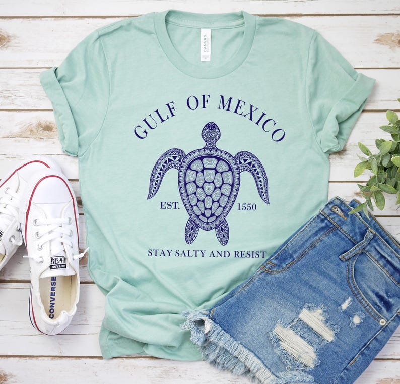Puede incluir: Una camiseta verde claro con un dise&ntilde;o de tortuga marina azul marino y el texto "GULF OF MEXICO" y "STAY SALTY AND RESIST". La camiseta se combina con pantalones cortos de mezclilla y zapatillas blancas.