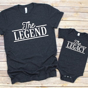 May include: A dark gray t-shirt with the text "The Legend" in white, and a dark gray baby onesie with the text "The Legacy" in white.