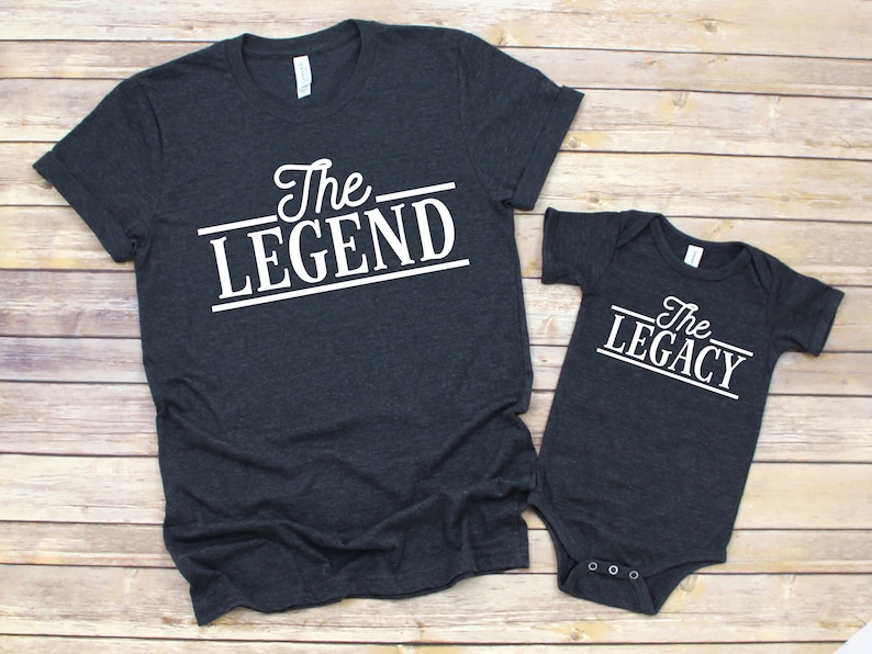 May include: A dark gray t-shirt with the text "The Legend" in white, and a dark gray baby onesie with the text "The Legacy" in white.