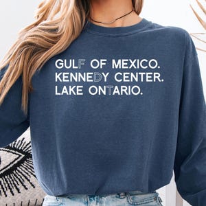 May include: A navy blue long-sleeve crewneck sweatshirt featuring white text that reads GULF OF MEXICO, KENNEDY CENTER, and LAKE ONTARIO. The letters F, D, and T are highlighted in a lighter shade within the words to spell out the acronym FDT.