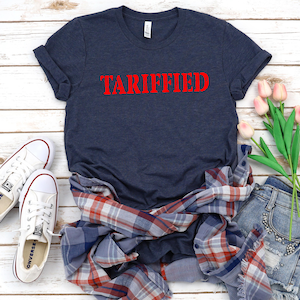 May include: A navy blue t-shirt with the word "TARIFFED" printed in red letters on the front. The shirt is laid out on a white wooden surface with a plaid shirt, a pair of jeans, and a bouquet of pink flowers.
