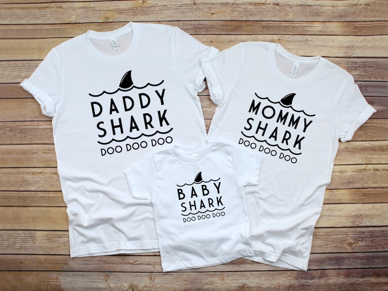 May include: Three white t-shirts with black text and a shark fin graphic. The shirts say "Daddy Shark Doo Doo Doo", "Mommy Shark Doo Doo Doo", and "Baby Shark Doo Doo Doo".