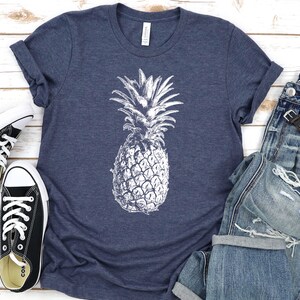 Pode incluir: Uma camiseta azul marinho com um gr&aacute;fico de abacaxi branco. A camiseta est&aacute; colocada sobre uma superf&iacute;cie de madeira branca com um par de sapatos de lona pretos e um par de cal&ccedil;as de ganga azuis com um rel&oacute;gio no bolso.
