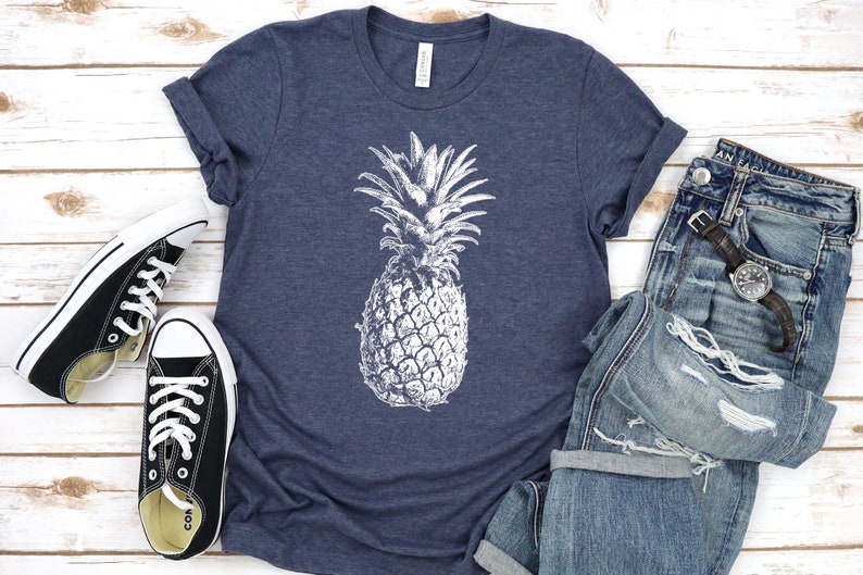 Pode incluir: Uma camiseta azul marinho com um gr&aacute;fico de abacaxi branco. A camiseta est&aacute; colocada sobre uma superf&iacute;cie de madeira branca com um par de sapatos de lona pretos e um par de cal&ccedil;as de ganga azuis com um rel&oacute;gio no bolso.