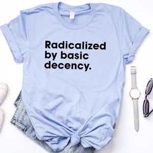 May include: A light blue t-shirt with the text "Radicalized by basic decency" in black. The shirt is styled with a pair of white shoes, denim shorts, a watch, and sunglasses on a white surface.