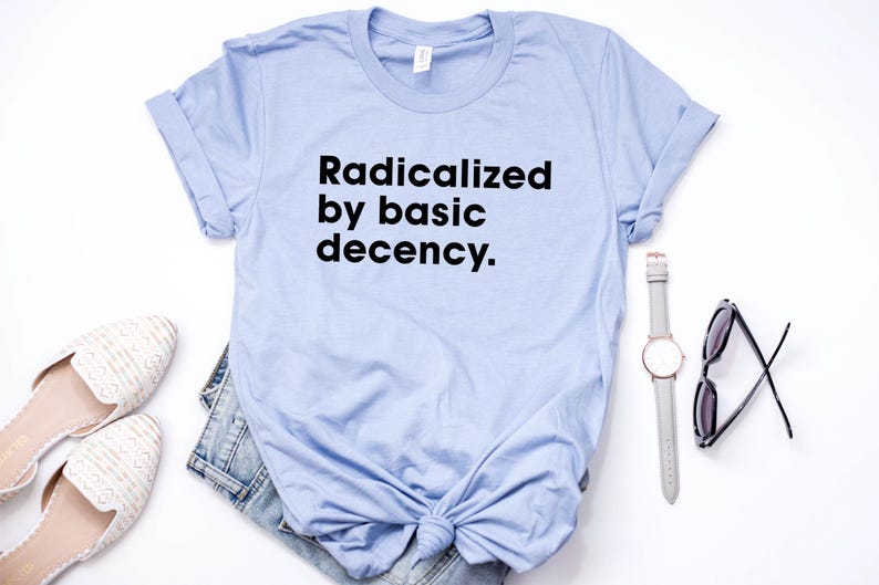 May include: A light blue t-shirt with the text "Radicalized by basic decency" in black. The shirt is styled with a pair of white shoes, denim shorts, a watch, and sunglasses on a white surface.