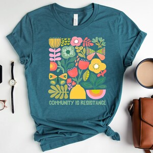 Op de afbeelding: Tealblauwe T-shirt met een kleurrijk bloemenontwerp en de tekst "Community is Resistance".